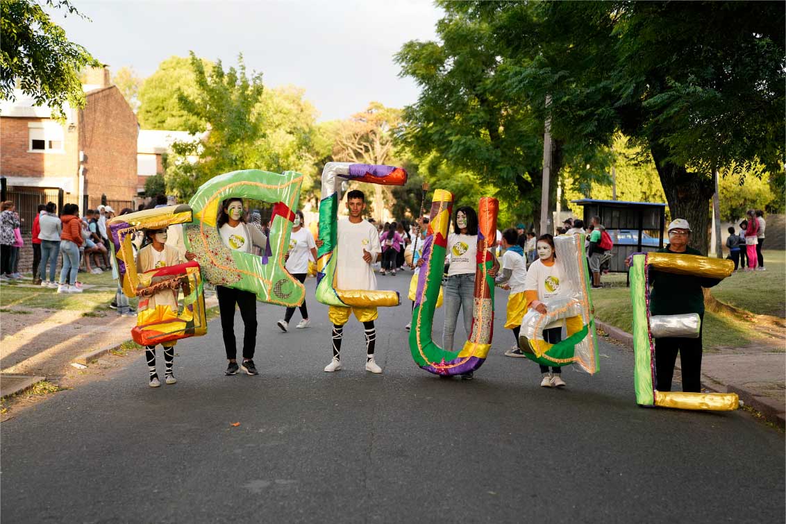 Cartel de Sacude sostenido por personas en un desfile. Letras de colores que son sostenidas cada una por personas distintas 