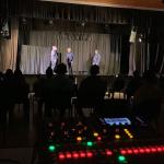 Se ven tres personas en un escenario durante una función en una sala con gente 