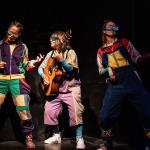 Fotografía de la obra La Mudanza, tres actrices en escena con ropa y lentes particulares, coloridos, una tiene una guitarra y las otras dos simulan tocar una.