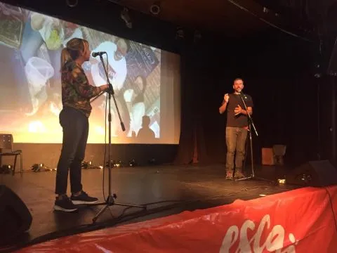 Una mujer y un hombre hablan sobre un escenario. Detrás se ve una fotografía proyectada.